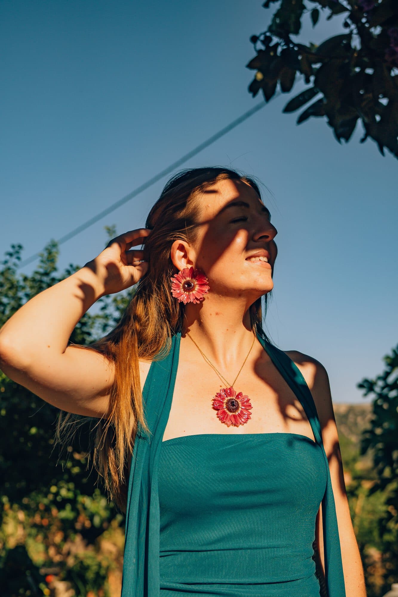 Modelo con pendientes y colgante de gerbera preservada de la Colección Invitada, a contraluz bajo un cielo azul